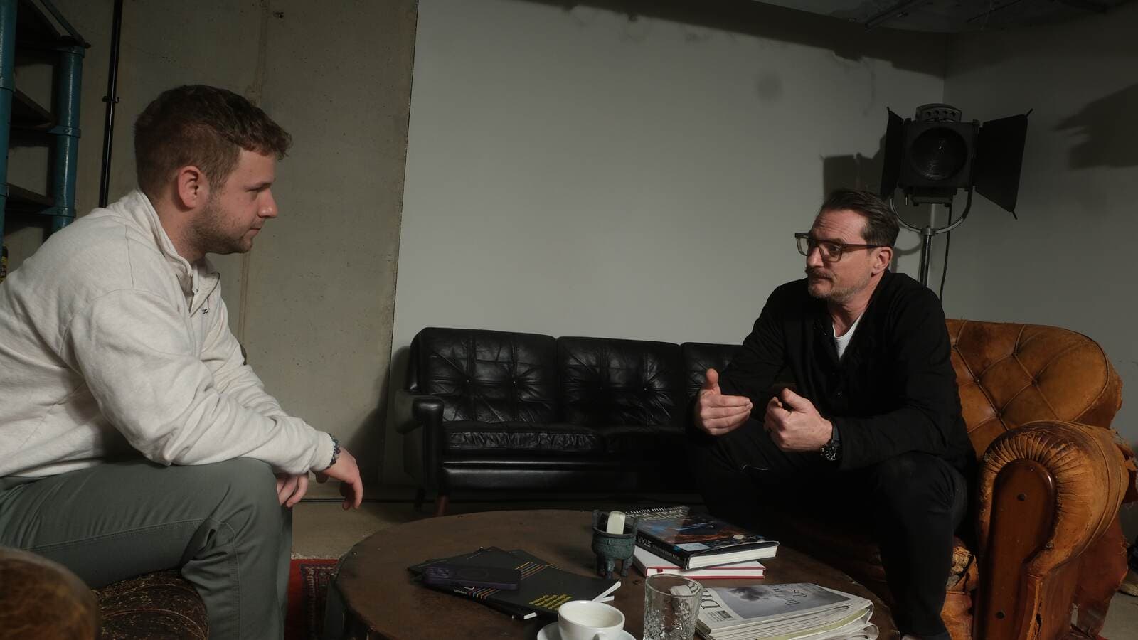 Manuel Kreutz im Gespräch mit Matteo Pieper; beide sitzen sich in Sesseln an einem niedrigen Tisch mit Büchern und Kaffeetasse gegenüber